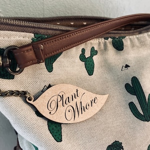 May include: A beige canvas bag with a brown leather strap and a wooden keychain with the text "Plant Whore" on it. The bag has a cactus print.