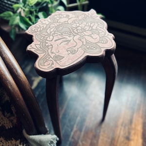 May include: A small, wooden plant stand with a Medusa face design carved into the top. The stand has three curved legs and a light brown finish. The top is approximately 12 inches in diameter.