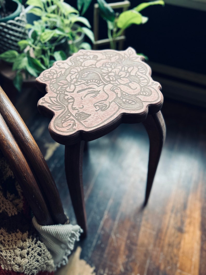 May include: A small, wooden plant stand with a Medusa face design carved into the top. The stand has three curved legs and a light brown finish. The top is approximately 12 inches in diameter.