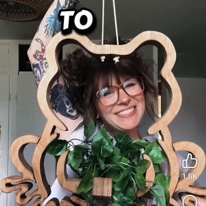 May include: A wooden frog-shaped wall hanging with a natural finish. The frog's outline is filled with green leafy plants. The word "TO" is visible at the top. The image has a light background.
