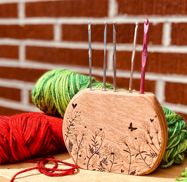 May include: Wooden stand with a floral design and a butterfly, holding five knitting needles and a crochet hook. The stand is surrounded by balls of yarn in red and green.