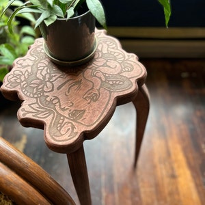 May include: A small, brown wooden plant stand with a carved Medusa design on the tabletop. The stand has three curved legs and holds a small potted plant with green leaves. The stand is approximately 12 inches tall.