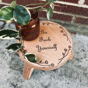 Pode incluir: Um pequeno suporte redondo de madeira para plantas com três pernas. O suporte tem a frase "So Fuck Yourself" gravada na madeira, rodeada por um desenho floral. Um vaso de vidro com uma planta está em cima.