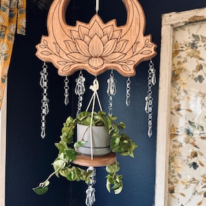 May include: A wooden plant hanger with a lotus flower and crescent moon design. Clear crystal beads hang below the wooden carving. A small gray pot with a green plant sits on a wooden shelf suspended below.