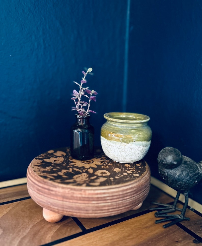 May include: A small wooden pedestal with a floral design, featuring a small glass bottle with purple flowers and a ceramic pot with a green and white glaze. A black metal bird figurine is on the right side of the pedestal.