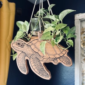May include: A wooden hanging planter shaped like a sea turtle, with a detailed engraved design. The planter holds a small green pot with a trailing green plant. White cords suspend the planter from above, creating a decorative and functional piece.