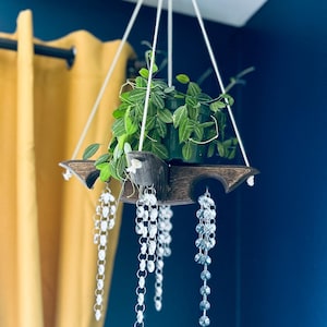 May include: A hanging wooden plant holder with a dark finish, suspended by white cords. The holder supports a potted plant with green foliage. Decorative crystal strands hang below, adding a touch of elegance. The background is a deep blue wall.