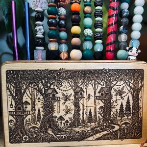 May include: A wooden box with a forest scene etched into the wood, holding several beaded pens and straws. The pens and straws have various colorful beads and charms, including a mushroom, bat, and owl. The text on the pen says "I like pretty things and the world fuck."
