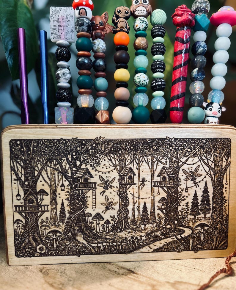 May include: A wooden box with a forest scene etched into the wood, holding several beaded pens and straws. The pens and straws have various colorful beads and charms, including a mushroom, bat, and owl. The text on the pen says "I like pretty things and the world fuck."