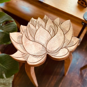 K&ouml;nnte beinhalten: Ein Hocker aus Holz mit einem geschnitzten Lotusbl&uuml;ten-Design. Der Hocker hat drei Beine und ist aus hellbraunem Holz gefertigt.