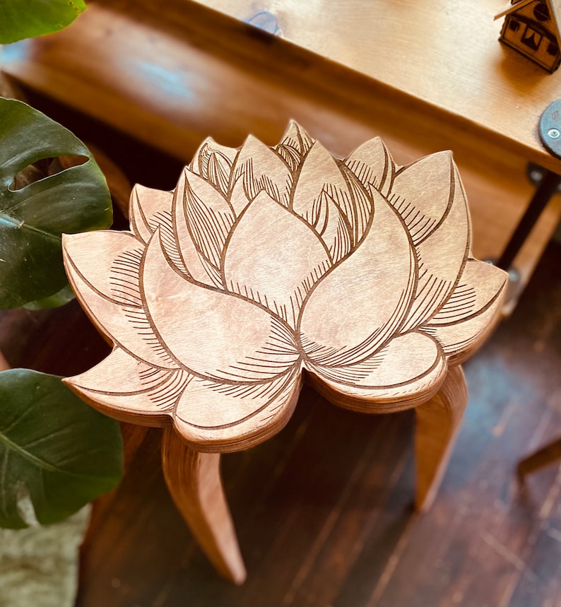K&ouml;nnte beinhalten: Ein Hocker aus Holz mit einem geschnitzten Lotusbl&uuml;ten-Design. Der Hocker hat drei Beine und ist aus hellbraunem Holz gefertigt.