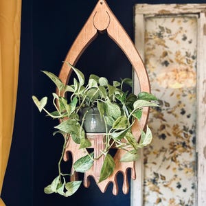 Puede incluir: Un colgador de plantas de madera con forma de lágrima y un diseño de goteo. El colgador está colgado del techo y tiene una planta en maceta dentro.