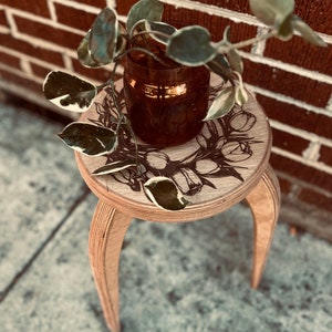 May include: A wooden stool with three legs and a round top. The top has a floral design and a brown glass vase with a leafy plant.