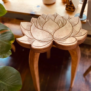K&ouml;nnte beinhalten: Ein Hocker aus Holz mit einem geschnitzten Lotusbl&uuml;tenmuster auf der Oberseite. Der Hocker hat drei gebogene und sich verj&uuml;ngende Beine.