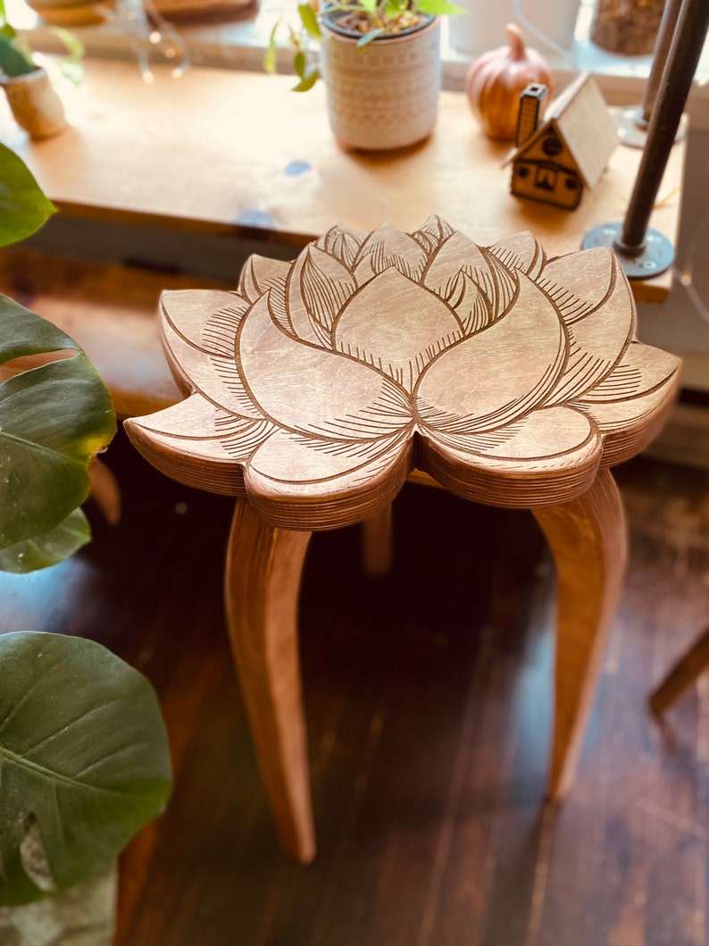 K&ouml;nnte beinhalten: Ein Hocker aus Holz mit einem geschnitzten Lotusbl&uuml;tenmuster auf der Oberseite. Der Hocker hat drei gebogene und sich verj&uuml;ngende Beine.