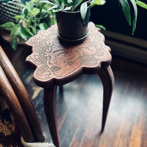 May include: A small, brown wooden plant stand with a carved face design on the tabletop. The stand has three curved legs and holds a dark gray pot with a green plant. The stand is approximately 12 inches tall.