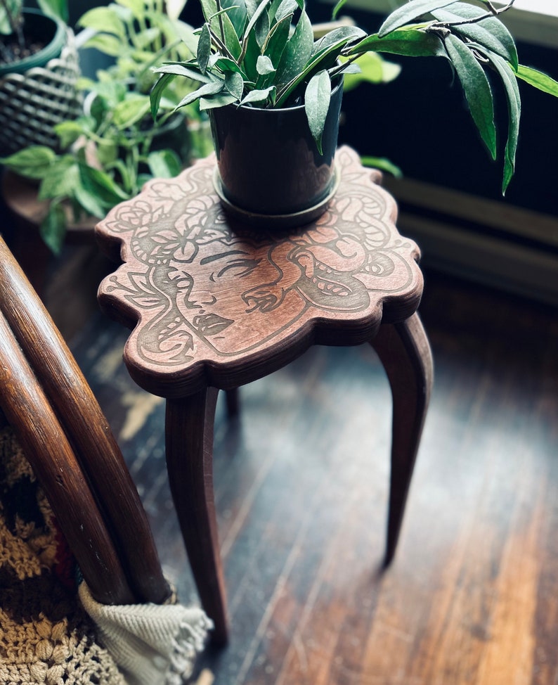 May include: A small, brown wooden plant stand with a carved face design on the tabletop. The stand has three curved legs and holds a dark gray pot with a green plant. The stand is approximately 12 inches tall.