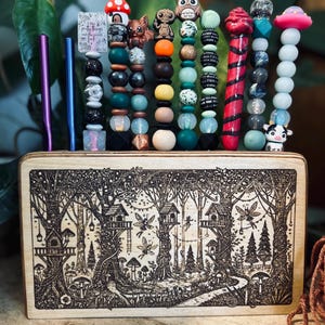 May include: A wooden organizer with a forest scene etched on the front, holding colorful beaded pens and straws. The pens and straws are purple and blue. The beads feature various shapes and characters, including an owl, a mushroom, and a cow.
