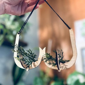 May include: A wooden hanging planter with two curved sections, designed to hold small plants. The planter is made of light brown wood and is hanging from a black string.