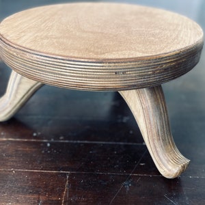 May include: A small, round wooden plant stand with three curved legs. The stand is made of light-colored wood with visible grain and a layered edge. The legs are a lighter shade of wood, providing a contrast to the top.