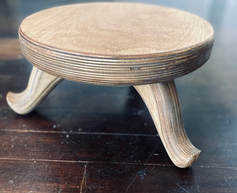 May include: A small, round wooden plant stand with three curved legs. The stand is made of light-colored wood with visible grain and a layered edge. The legs are a lighter shade of wood, providing a contrast to the top.