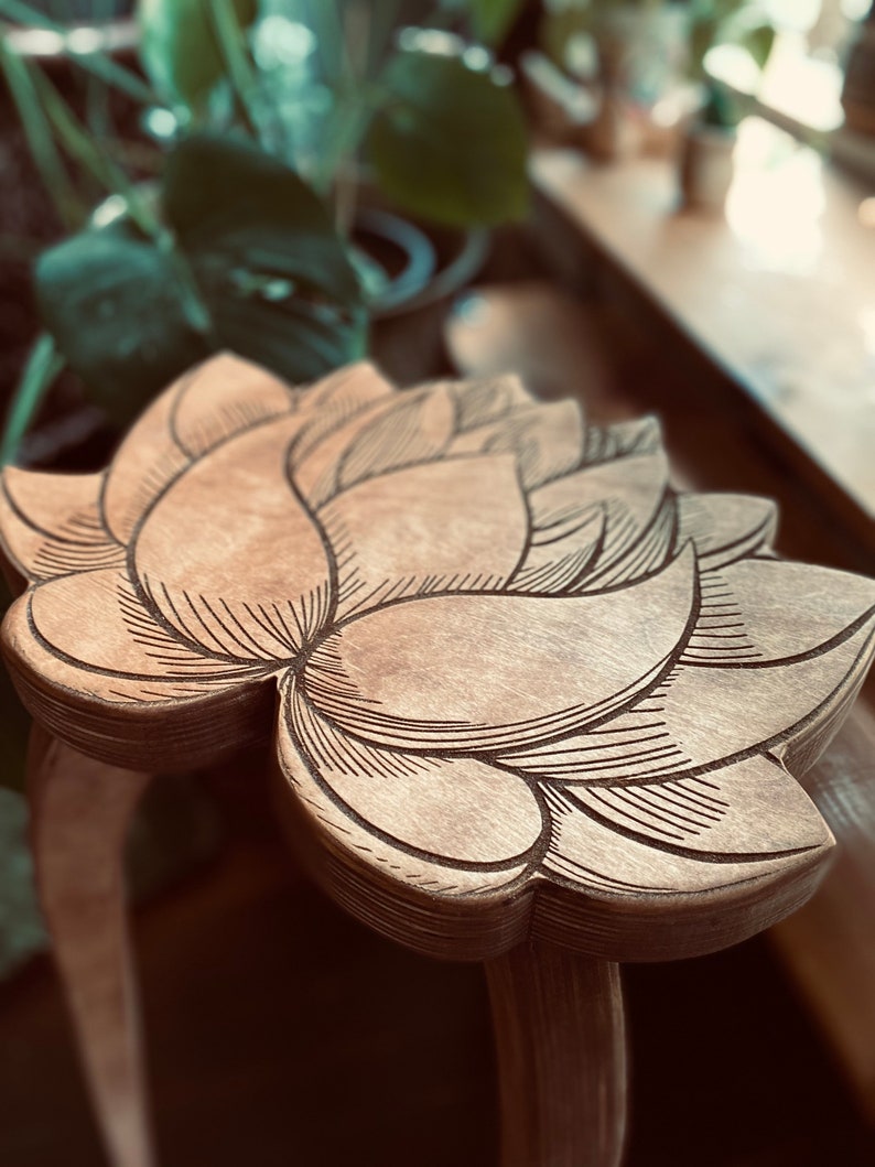 K&ouml;nnte beinhalten: Ein Hocker aus Holz mit einem geschnitzten Lotusblumenmuster auf der Oberseite. Der Hocker ist aus hellbraunem Holz gefertigt und hat eine nat&uuml;rliche Oberfl&auml;che.