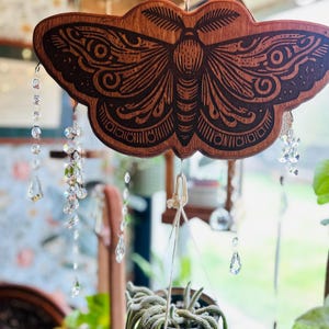 May include: A wooden cutout of a moth with intricate details hangs from a string with crystal beads. The moth is brown with black details and is suspended in front of a window.