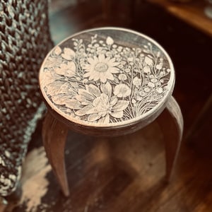 May include: A wooden stool with a carved floral design. The design features white flowers and leaves on a brown background.