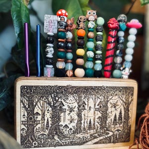 May include: A wooden box with a forest scene etched on the front, holding several beaded pens and two metal pens. The beaded pens feature various charms, including an owl, mushroom, and alien. One pen has the text "I like pretty things and the good luck."