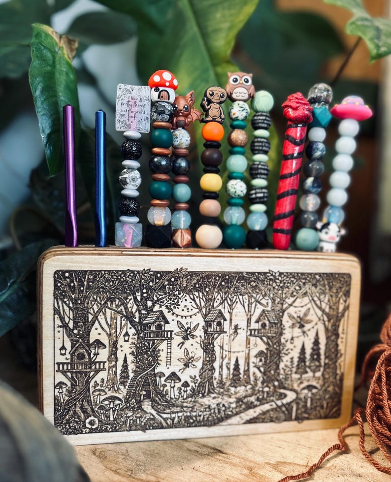 May include: A wooden box with a forest scene etched on the front, holding several beaded pens and two metal pens. The beaded pens feature various charms, including an owl, mushroom, and alien. One pen has the text "I like pretty things and the good luck."