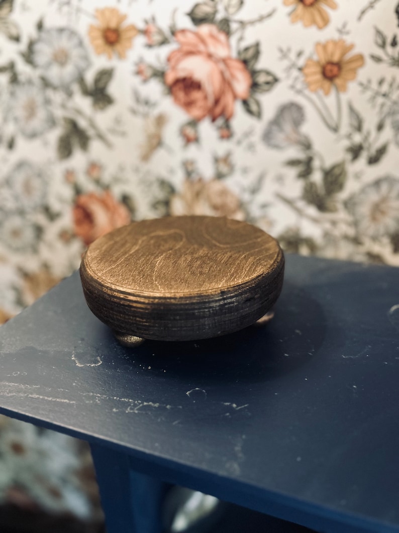 May include: A round wooden pedestal with a gold finish and three legs. The pedestal is sitting on a blue surface.