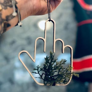 May include: A wooden ornament in the shape of a hand giving the middle finger, with a small evergreen sprig inside. The ornament is suspended by a black string.