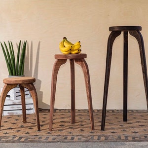 May include: Three wooden plant stands of varying heights. The stands have a tripod leg design and circular tops. The shortest stand holds a potted plant, the middle stand holds a bunch of bananas, and the tallest stand is empty. The wood tones vary from light to dark.