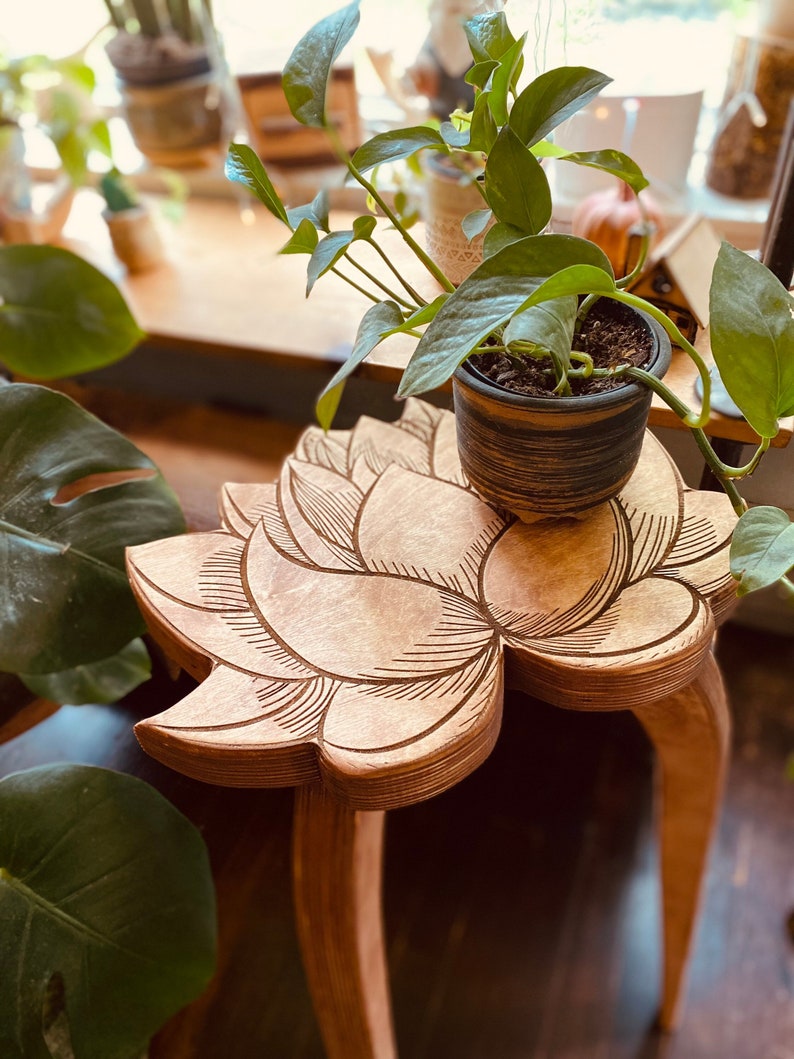 K&ouml;nnte beinhalten: Ein Holz-Beistelltisch mit einem geschnitzten Lotusblumen-Design. Der Tisch hat drei Beine und ist mit einer Topfpflanze dekoriert.
