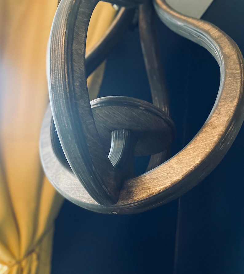 May include: A close-up of a wooden, dark brown, decorative hanging ornament with a knot-like design. The ornament is made of multiple pieces of wood that are intricately woven together.