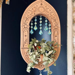 May include: A hanging wooden planter with an arched frame featuring laser-engraved celestial motifs including stars and moon phases. The frame holds a small green plant and is decorated with five dangling iridescent blue crystal prisms.