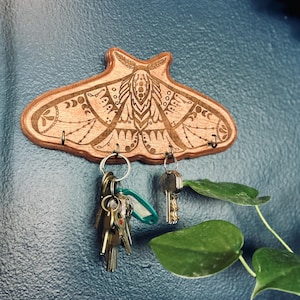 May include: Wooden key holder shaped like a moth, with intricate engraved details. The key holder has four hooks and is holding several keys. The key holder is mounted on a textured blue wall. A few green leaves are in the background.