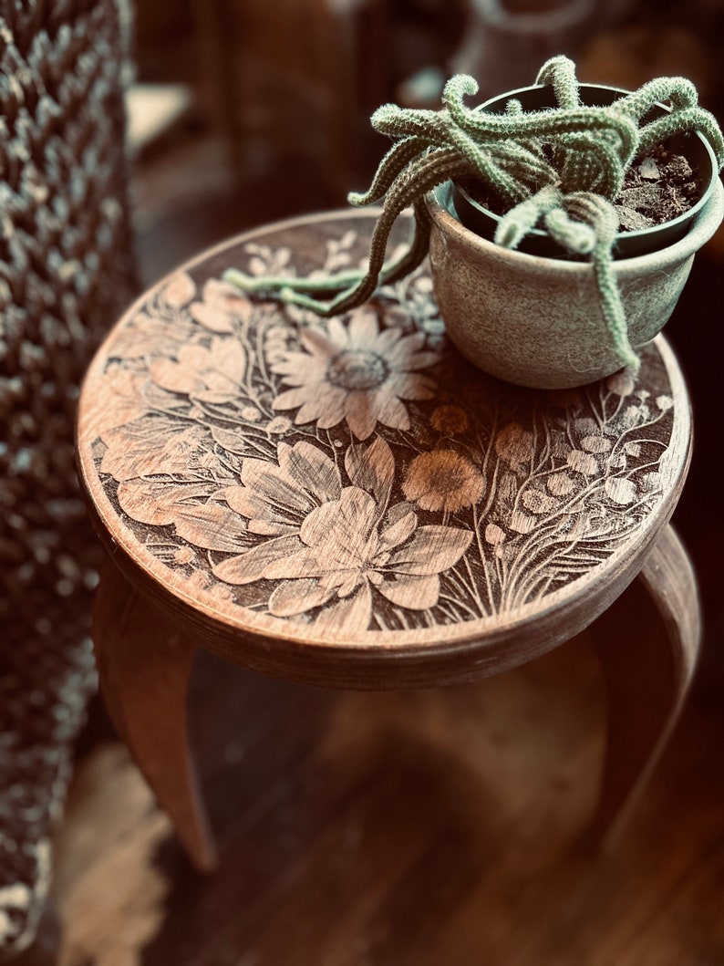 May include: A wooden stool with a floral design carved into the top. A small potted cactus plant sits on top of the stool.