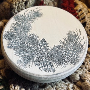May include: A round wooden coaster with a black and white design of pine branches and pine cones.