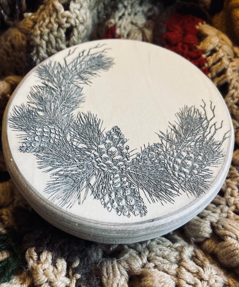 May include: A round wooden coaster with a black and white design of pine branches and pine cones.
