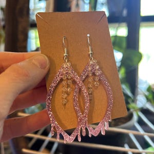 Peut inclure: Boucles d'oreilles roses en forme de goutte d'eau avec des paillettes et un motif dégoulinant. Les boucles d'oreilles ont un crochet argenté et un groupe de perles transparentes suspendues au centre. Présentées sur une carte marron.