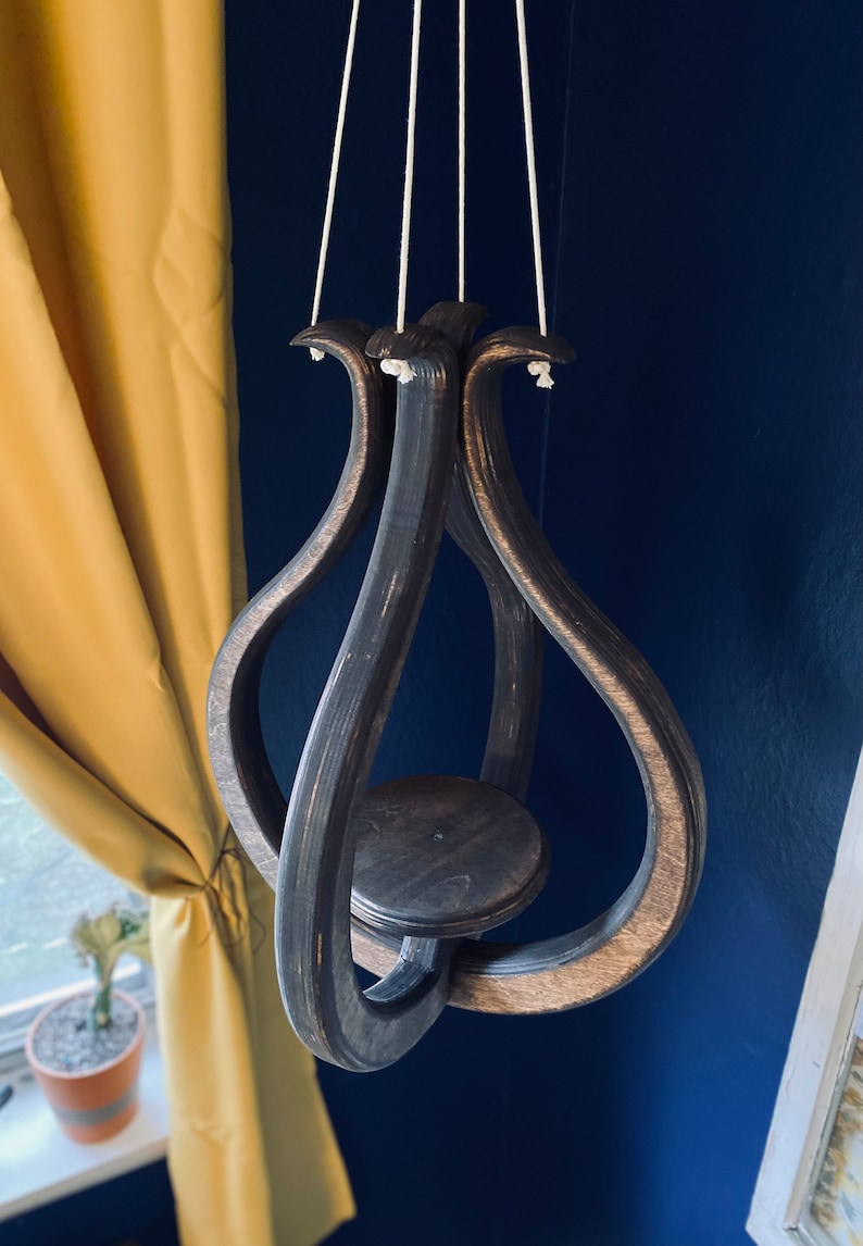 May include: A dark brown wooden hanging planter with a circular base and a unique, intertwined design. The planter is suspended by three white cords.