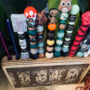 May include: A collection of beaded crochet hook handles with various decorative toppers, including a mushroom, a dragon, and an owl. The handles are displayed in a wooden holder with a forest scene. Some handles have text that reads "I like pretty things and the word f*ck".
