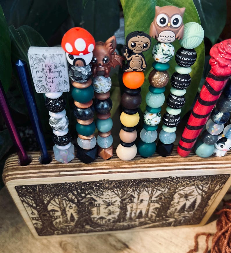 May include: A collection of beaded crochet hook handles with various decorative toppers, including a mushroom, a dragon, and an owl. The handles are displayed in a wooden holder with a forest scene. Some handles have text that reads "I like pretty things and the word f*ck".