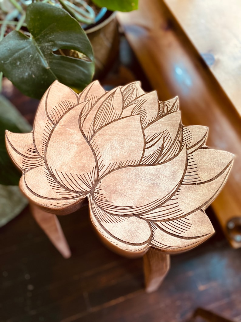 K&ouml;nnte beinhalten: Ein Hocker aus Holz in Form einer Lotusblume mit aufwendigen Details, die in die Oberfl&auml;che geschnitzt sind. Der Hocker ist aus hellbraunem Holz gefertigt und hat eine nat&uuml;rliche Oberfl&auml;che.