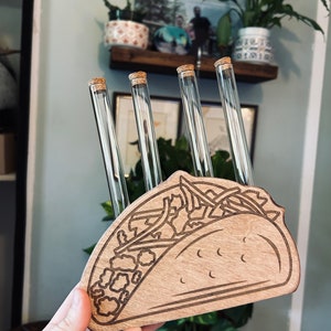 May include: Wooden taco-shaped holder with four glass test tubes with cork tops. The holder is engraved with a detailed outline of a taco with fillings.