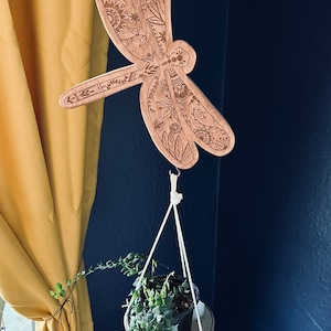 Puede incluir: Una libélula de madera con intrincados diseños cortados con láser cuelga de un colgador de plantas de macramé. Una pequeña planta en maceta con enredaderas colgantes cuelga debajo de la libélula.