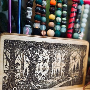 May include: A wooden organizer with a forest scene etched on the front, holding colorful beaded pens and accessories. The pens feature various beads in shades of green, brown, and red, with whimsical charms.