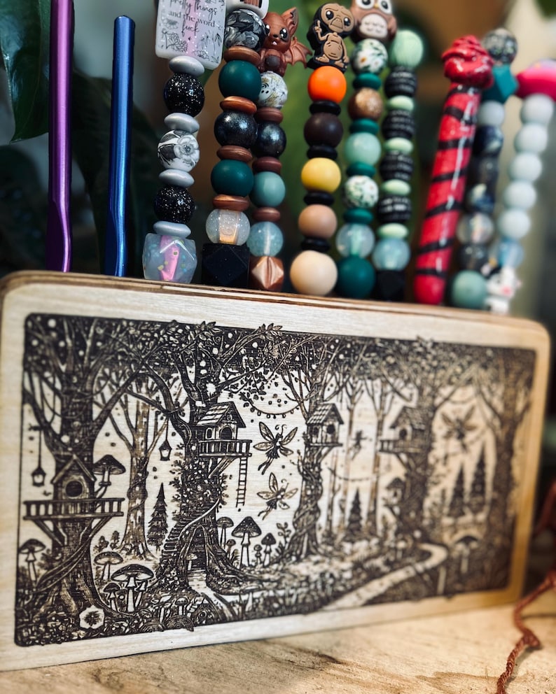 May include: A wooden organizer with a forest scene etched on the front, holding colorful beaded pens and accessories. The pens feature various beads in shades of green, brown, and red, with whimsical charms.