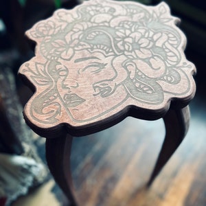 May include: A small, wooden side table with a Medusa design carved into the tabletop. The table has three curved legs and a dark brown finish. The Medusa design features a woman's face with snakes and floral elements.